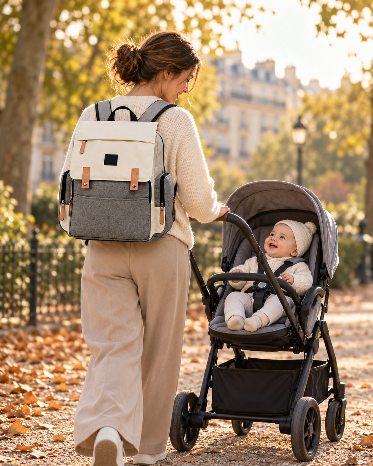Une mère porte le sac à langer Land beige sur le dos et pousse son bébé souriant dans un parc parisien, en automne