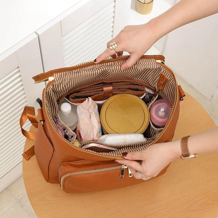 Deux mains ouvrent un sac à dos à langer en cuir caramel rempli d’affaires de bébé bien rangées