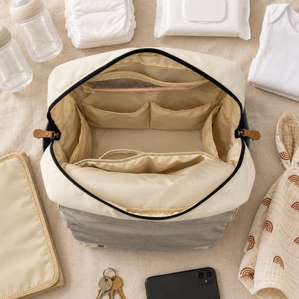 Le sac Land grand ouvert, avec ses poches intérieures, entouré de biberons, de couches, du tapis à langer et d’un doudou