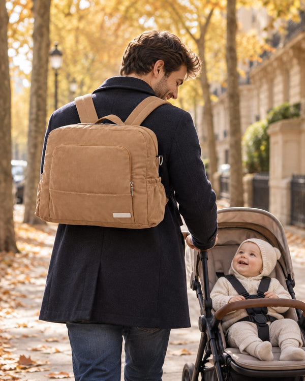Un père porte un sac à dos à langer en velours côtelé caramel et pousse son bébé souriant dans une avenue parisienne, en automne