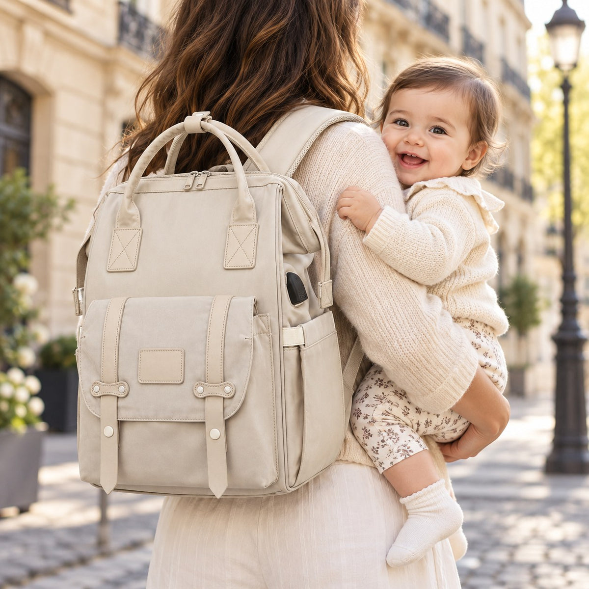 Sac à dos à langer beige avec port USB porté sur le dos, bébé souriant dans les bras de son parent